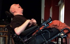 Here is a picture of me, reclining back in my wheelchair laughing hard, while wearing a clean shave, a black tee, and orange corduroy pants.